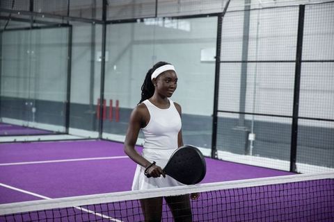 African american woman holding padel racket at net on purple indoor court in white dress
