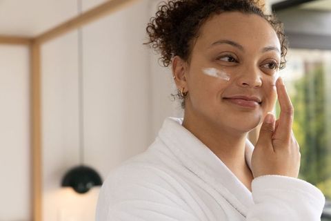 African american woman applying facial moisturizer by bedroom window wearing white robe