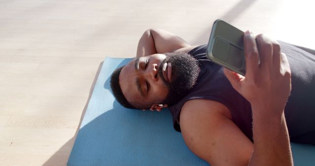 African American man lying on exercise mat uses smartphone in indoor setting. Man appears relaxed and engaged with his phone, illustrating technology use in leisure time. Suitable for themes involving relaxation, fitness, technology integration in daily life, exercise breaks.