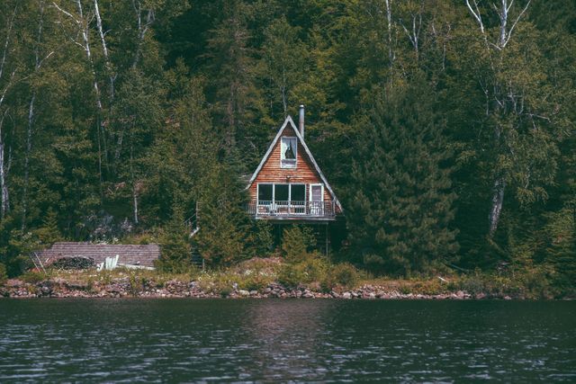 A rustic A-frame cabin nestled in dense forest by a calm lake, with lush trees surrounding the water. This image can be used for promoting nature retreats, holiday getaways, outdoor activities, and real estate listings for houses in scenic locations.