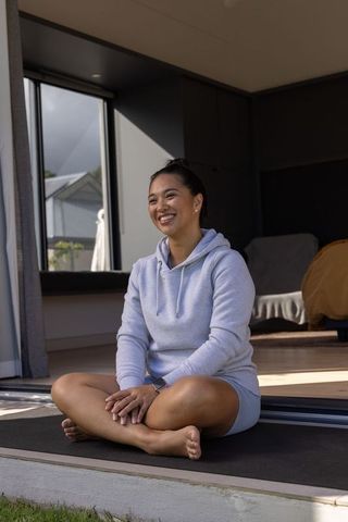 Smiling asian woman sitting cross-legged on mat at open doorway wearing gray hoodie and smartwatch