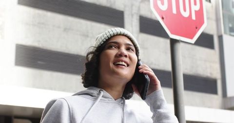 Smiling young woman wearing beanie talking on smartphone on urban sidewalk near stop sign