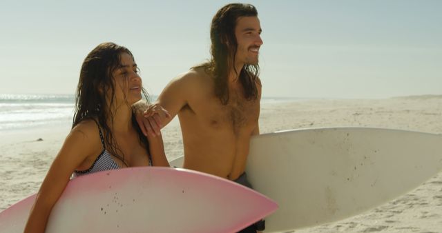 Young Caucasian man and biracial woman hold surfboards on the beach. They share a moment of joy in the sun, ready for a surf session outdoors.
