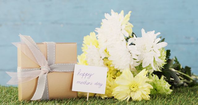 A gift box tied with a ribbon and a bouquet of yellow and white flowers are accompanied by a 'Happy Mother's Day' note, with copy space. These items symbolize appreciation and celebration for Mother's Day.