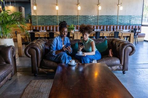 African american friends relaxing on leather sofa scrolling smartphones in urban cafe