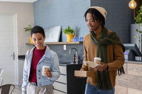 Diverse friends walking through modern coworking kitchenette carrying coffee cups and takeaway box