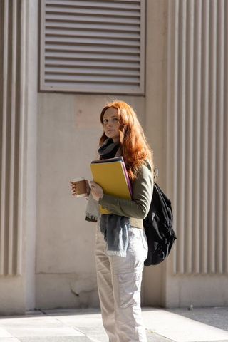 Red-haired college student standing with colorful folders and coffee by campus columns