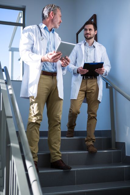 Two doctors in lab coats are walking down a hospital staircase while discussing something on their tablets. This image can be used to depict teamwork and communication among medical professionals, modern healthcare settings, and the use of technology in medical environments.