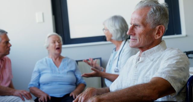 Senior individuals engage in a discussion or activity in a room, a care facility, with a nurse or facilitator present. The setting suggests a social or therapeutic gathering aimed at promoting engagement and well-being among the elderly.