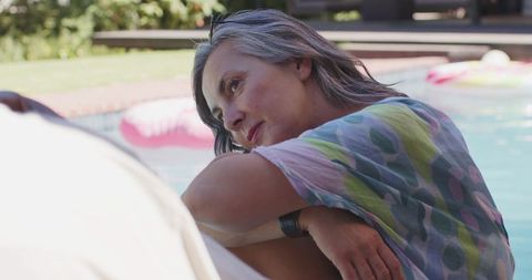 Mature woman relaxing at backyard pool edge wearing patterned coverup and sunglasses