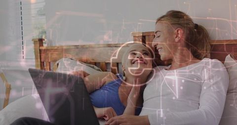 Female couple sharing laptop on bed with holographic tech overlay, cozy intimate moment