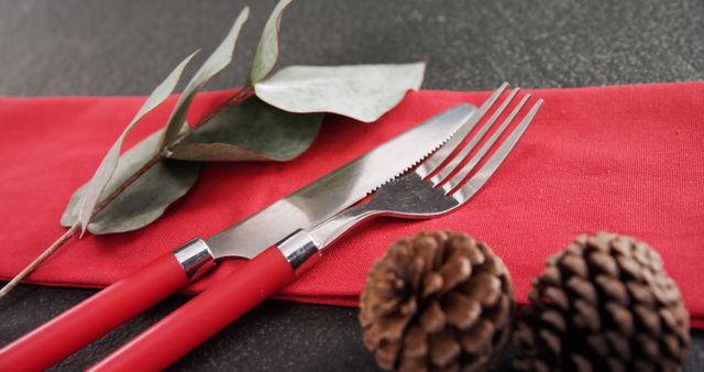 A festive table setting features a knife and fork with red handles on a napkin, accompanied by pine cones and eucalyptus leaves, with copy space. The arrangement suggests a holiday meal or a celebratory event, inviting a sense of warmth and tradition.