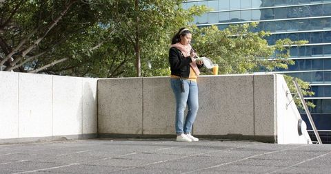 Asian woman standing on urban plaza holding smartphone and takeaway coffee, casual break