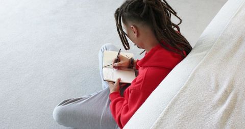 Mid-adult man with dreadlocks writing in notebook on minimal grey carpet cozy journaling