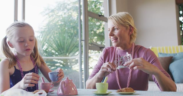 Grandmother and granddaughter having a tea party together while wearing pink beads. This heartwarming scene captures family bonding, playtime, and shared joy. Ideal for concepts related to family relationships, childhood activities, indoor fun, and intergenerational connections.