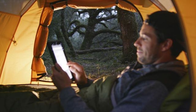 Adult man using tablet in yellow tent at twilight forest camping relaxation cozy outdoors