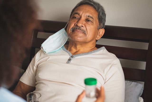 Senior man wearing face mask lying in bed smiling during a home medical visit. Ideal for use in healthcare, elderly care, and COVID-19 related content. Can be used in articles, blogs, and advertisements focusing on home healthcare services, senior care, and pandemic safety measures.