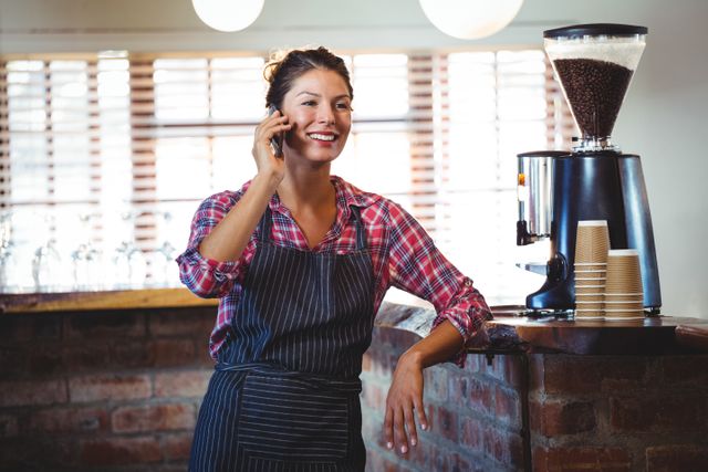 Waitress in casual attire making a phone call in a cozy cafe. Ideal for use in content related to customer service, hospitality industry, small businesses, and communication. Can be used in articles, blogs, and advertisements focusing on cafes, baristas, and friendly service environments.