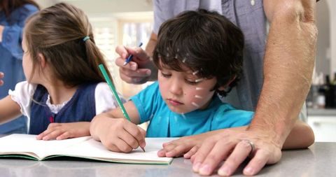 Father guiding young boy writing at kitchen counter with gentle adhesive strips on face