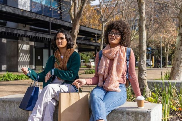Two women sitting on urban bench holding shopping bags and coffee, chatting outdoors