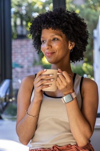 African woman holding ceramic mug, wearing smartwatch, smiling, relaxing in sunlit sunroom
