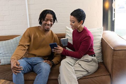 African american couple sharing smartphone and coffee while relaxing on cozy leather sofa