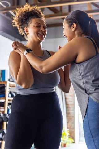 Diverse women smiling and holding forearms during supportive gym training session