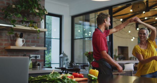 A couple is dancing joyfully in a contemporary kitchen setting while preparing food together. They are surrounded by fresh vegetables, including bell peppers and greens. The kitchen features large windows, brick walls, and indoor plants, giving a cozy and modern vibe. Ideal for use in promotional materials for kitchen appliances, cooking classes, or lifestyle blogs focusing on relationships and home decor.