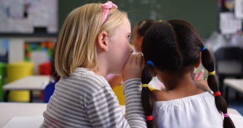 Two young students whispering with colorful hair ties in classroom setting