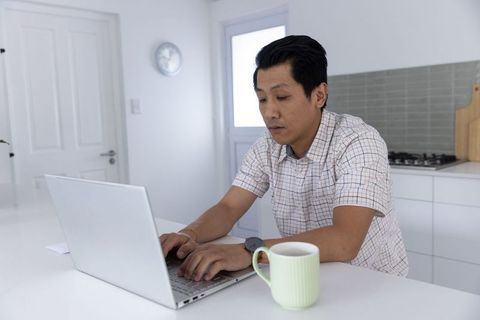 Asian man working on laptop at minimalist kitchen counter with coffee mug for remote work