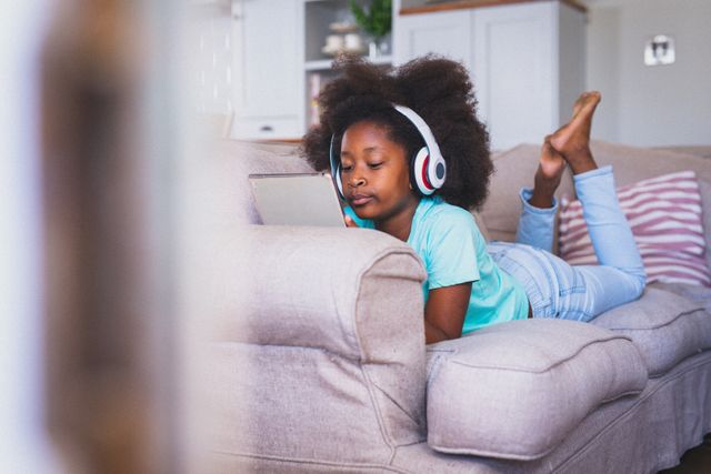 African american girl wearing headphones lying on couch using tablet. staying at home in isolation during quarantine lockdown.