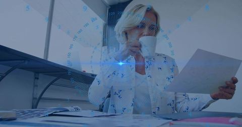 Businesswoman reviewing documents while sipping coffee with holographic overlay at desk