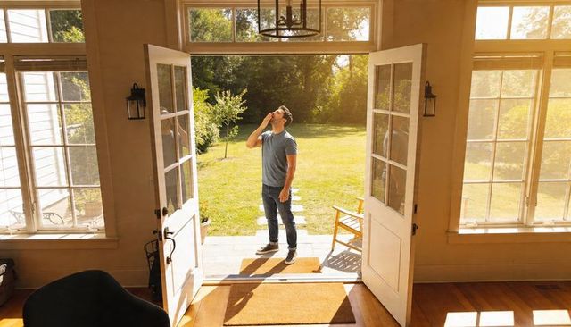 Young man standing on tiled step at open front doors holding phone in sunlit suburban entryway