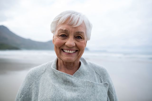 Senior woman smiling while standing on beach, perfect for promoting healthy lifestyle, retirement plans, travel destinations, and senior wellness programs. Ideal for use in advertisements, brochures, and websites targeting older adults.