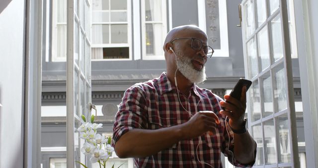 Elderly man in casual plaid shirt wearing glasses and using smartphone with headphones in bright, airy room. Perfect for themes of technology use among seniors, relaxation, staying connected, enjoying modern gadgets, indoor lifestyle, and casual living. Useful for articles, blog posts, advertisements, and social media campaigns focusing on senior experiences with technology or music enjoyment.