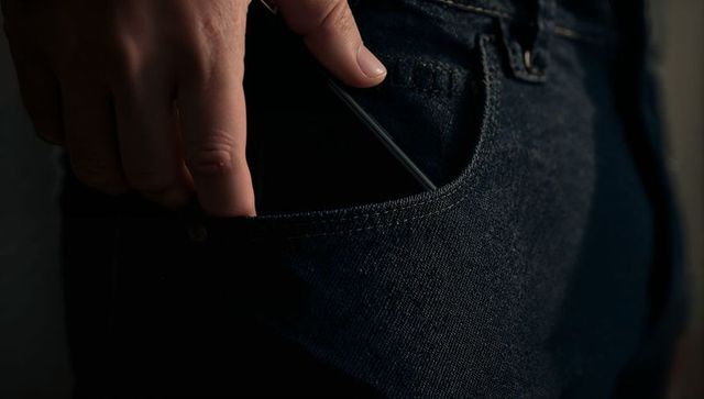 Hand sliding smartphone into dark denim front pocket closeup moody low-light texture grainy