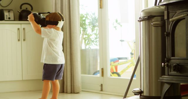 Young boy explores technology by trying a virtual reality headset in a bright kitchen. Modern home environment enhances the emphasis on innovation and curiosity. Ideal for technology blogs, child development articles, and advertisements featuring modern gadgets.