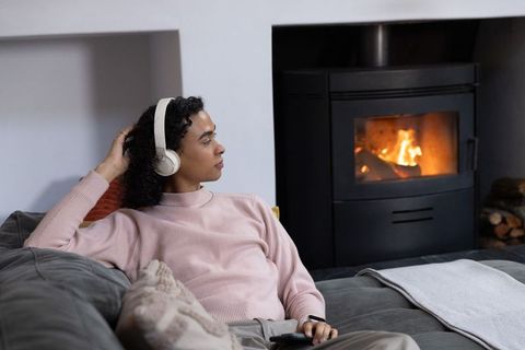 African american nonbinary person relaxing on couch with over-ear headphones by fireplace