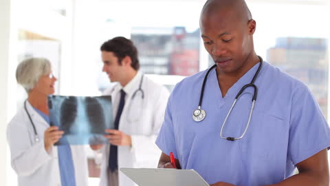 Healthcare Professional Documenting While Colleagues Consult in Background