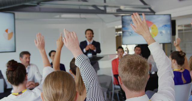 Professional gathering showing active participation of diverse employees raising hands during a business presentation. This setting is excellent for illustrating corporate training sessions, teamwork, employee engagement, and collaboration in a professional context. Ideal for use in business resources, educational materials, and team-building manuals.