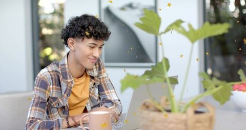 Young man working remotely on laptop at bright home patio with plants and coffee