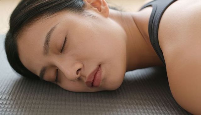 Asian woman resting on grey yoga mat, eyes closed, embracing quiet relaxation