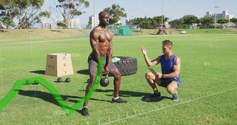 Shirtless athlete swinging kettlebell while coach instructing outdoor strength session
