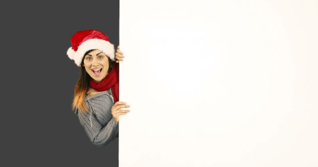 An excited woman wearing a Santa hat, grey sweater, and red scarf is peeking from behind a blank sign board, looking surprised and happy. Perfect for Christmas and holiday season advertisements, festive greetings, and seasonal marketing materials.