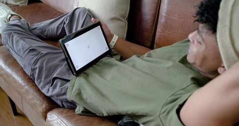 African american young man reclining on leather sofa holding tablet and relaxing at home