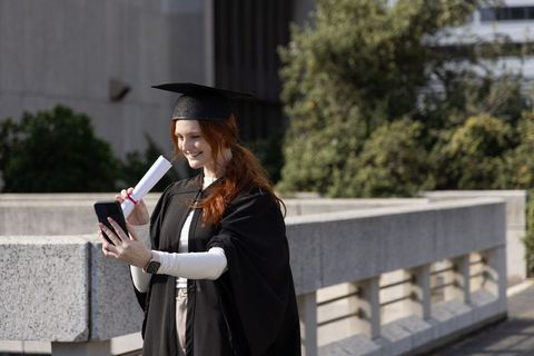 Graduate taking selfie on campus terrace wearing gown and mortarboard holding diploma
