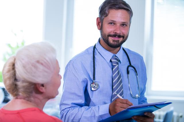 This image shows a friendly doctor consulting with an elderly patient in a hospital setting. The doctor is smiling and holding a clipboard, suggesting a positive and reassuring consultation. Stethoscope around the doctor's neck indicates a medical professional. This image is ideal for use in healthcare articles, medical blogs, senior care websites, and health service promotions.
