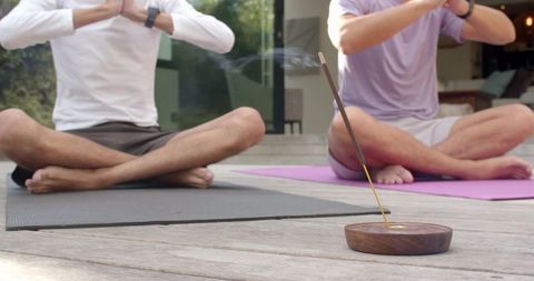 Two men practicing outdoor meditation on wooden deck with burning incense and yoga mats