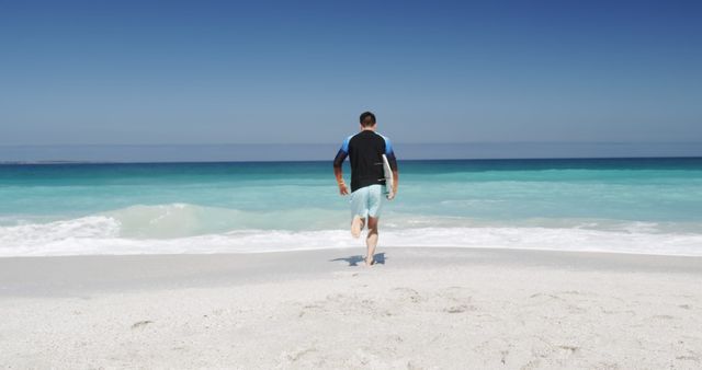 Man walking along sandy beach towards ocean with clear blue sky. Ideal for travel and tourism, summer vacation promotions, relaxation, leisure activities, and nature conservation themes.
