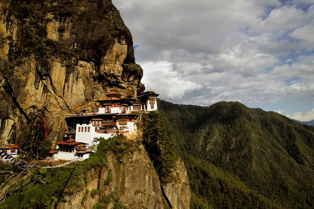 Tiger's Nest Monastery nestled on a steep cliff edge in Bhutan. Wooden structures and intricate Bhutanese architecture are evident. Vast forested mountains spread in the background beneath a partly cloudy sky. Ideal for travel blogs, cultural exploration articles, and landscape photography showcases. Perfect for highlighting exotic travel destinations and historical architecture.